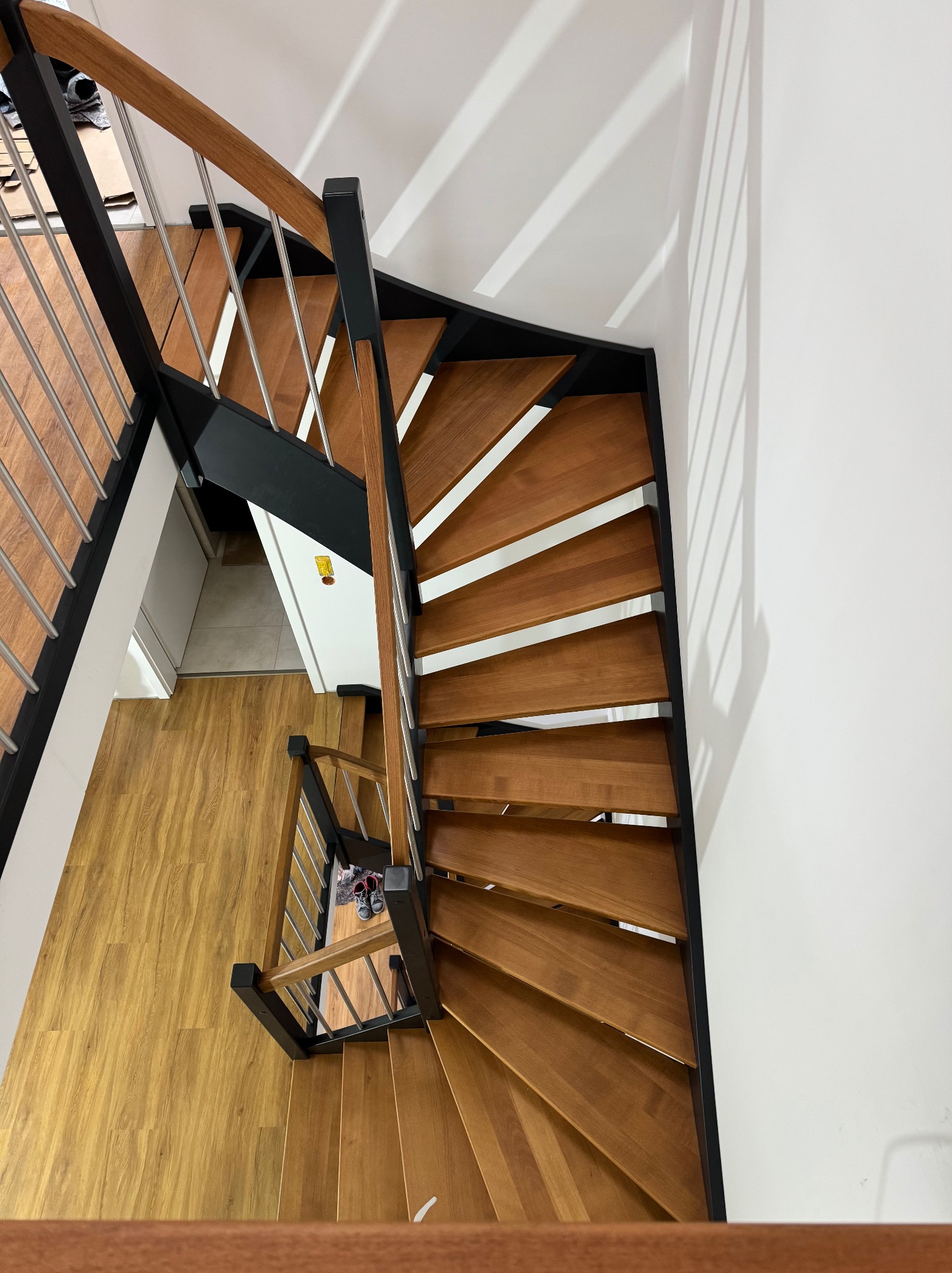 Widok z góry na drewniane schody z czarną metalową konstrukcją i srebrnymi prętami balustrady, oświetlone naturalnym światłem padającym na białą ścianę obok.