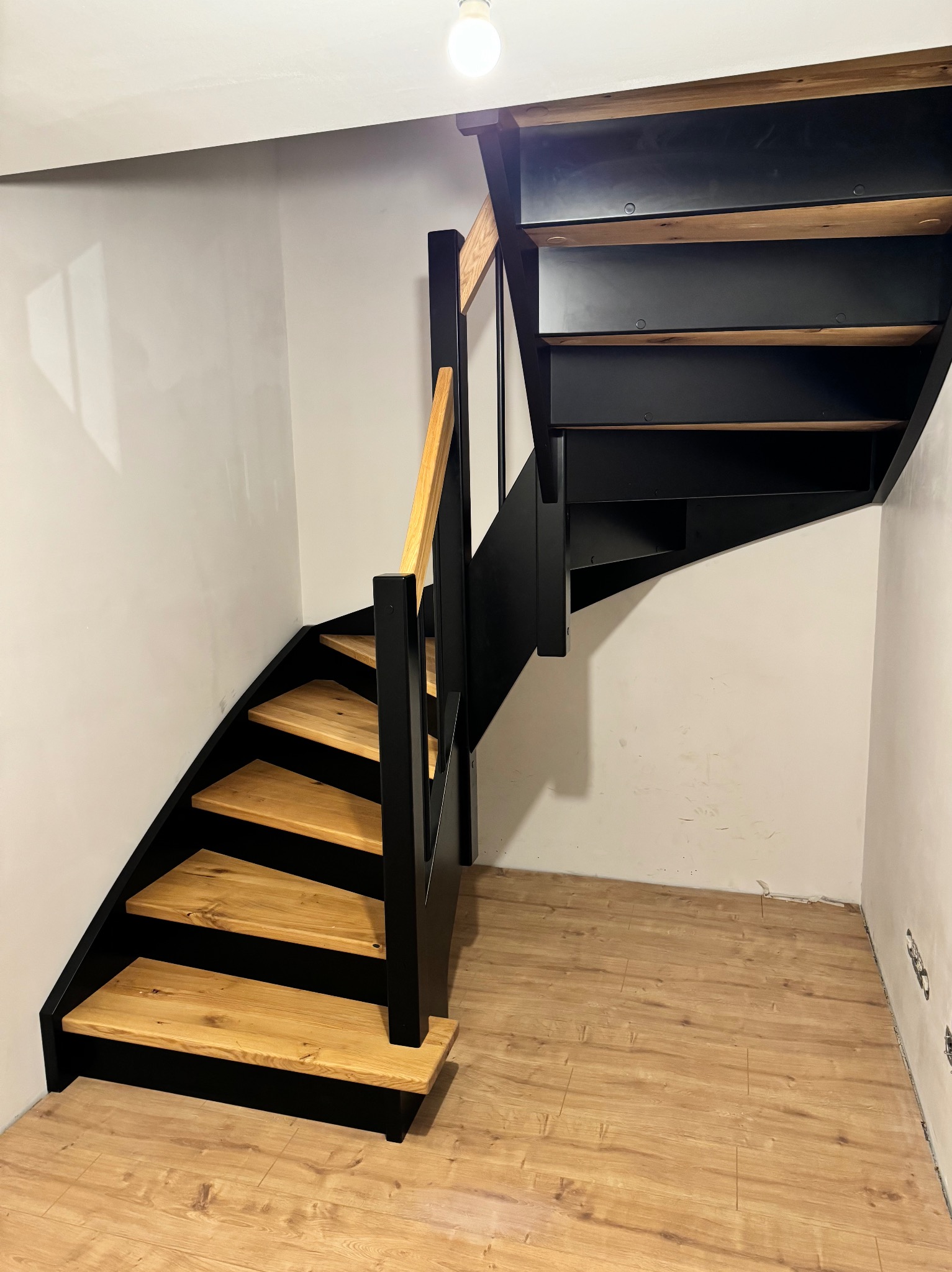 Nowoczesne schody z drewnianymi stopniami i czarną metalową konstrukcją, widoczne od dołu, z drewnianą poręczą, na tle białych ścian i drewnianej podłogi.