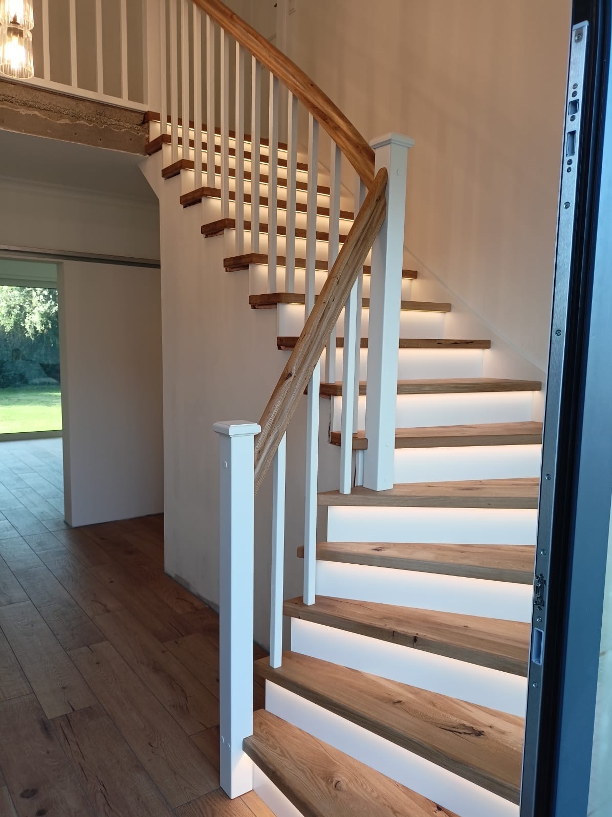 Nowoczesne schody z drewnianymi stopniami podświetlonymi od spodu, białe balustrady i drewniana poręcz, widok z dołu na jasne wnętrze.