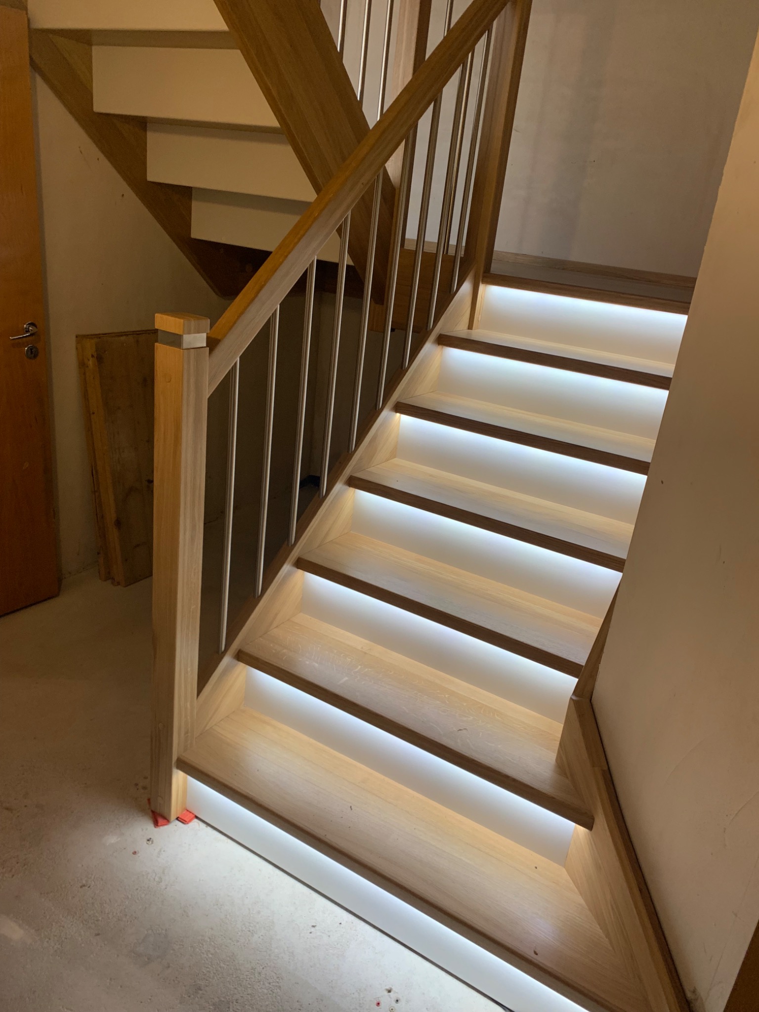 Jasne, drewniane stopnie schodów z podświetleniem LED, zabezpieczone metalową balustradą z pionowymi prętami, widoczne w trakcie montażu, z podkładkami poziomującymi u podstawy.