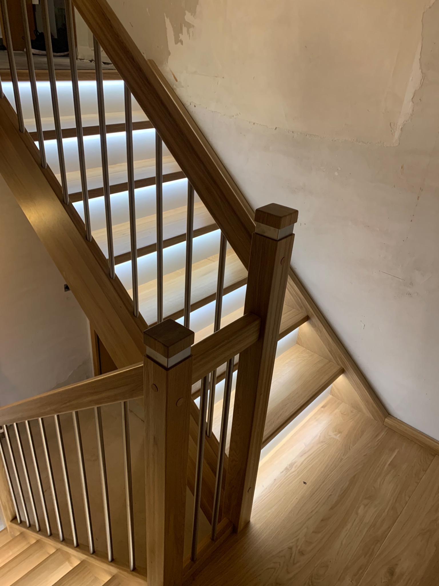 Jasne, drewniane schody z metalowymi prętami balustrady i podświetleniem LED stopni, widoczne niedokończone ściany w tle.