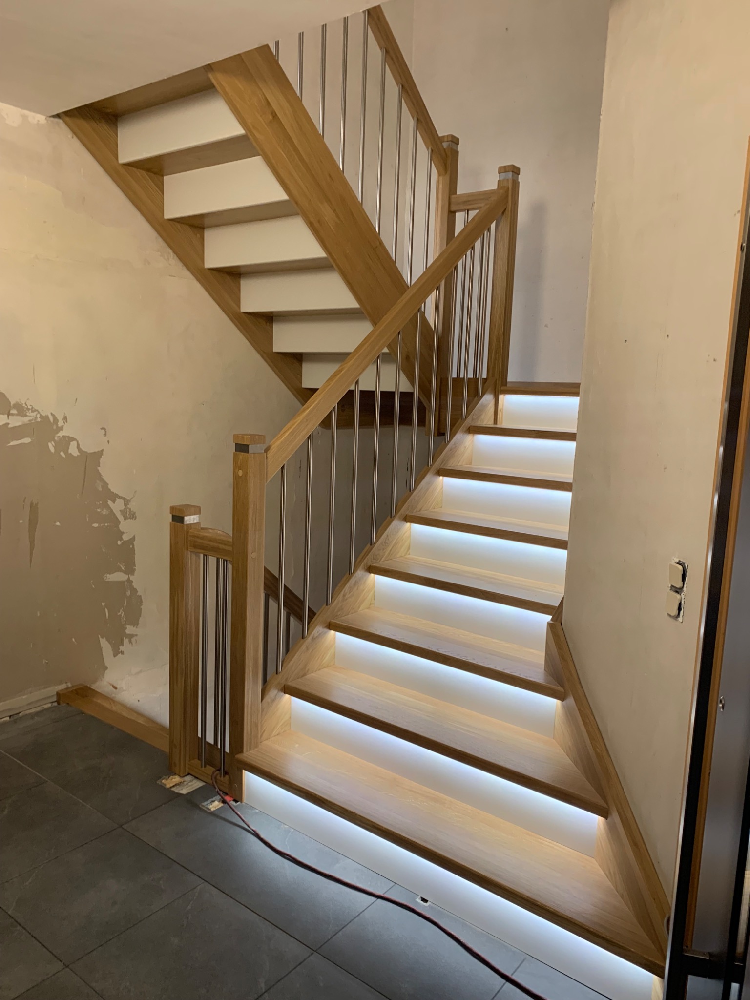 Drewniane schody z podświetleniem LED na stopniach i metalowymi poręczami, widoczne niedokończone wykończenie ściany i podłogi.