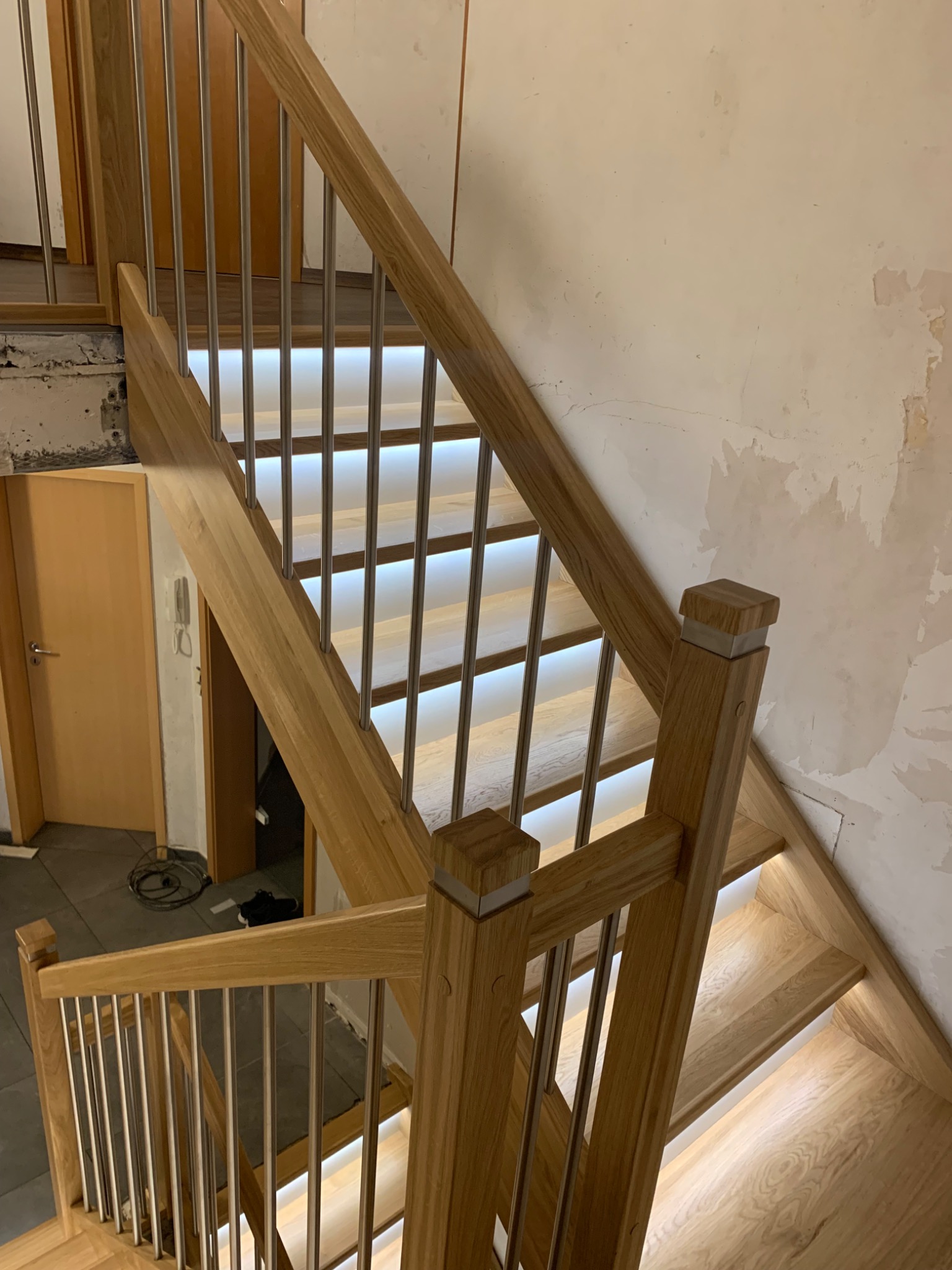 Jasne drewniane schody z podświetleniem LED stopni i metalowymi poręczami w trakcie montażu, widoczne niedokończone ściany i podłogi.