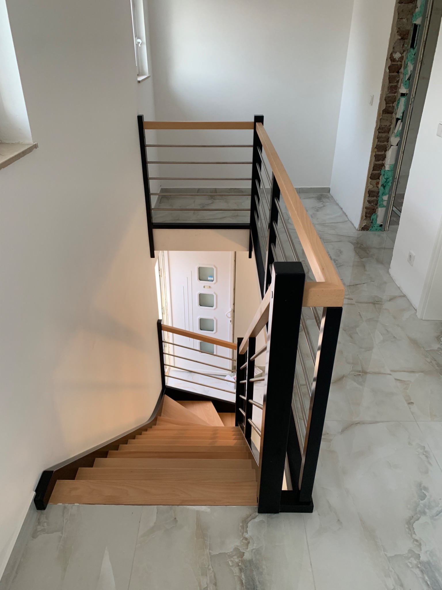 Nowoczesne schody drewniane z czarnymi słupkami i metalowymi prętami balustrady, widok z góry na dół, z marmurową podłogą i białymi ścianami, fragment wykończenia ściany bez tynku po prawej stronie.