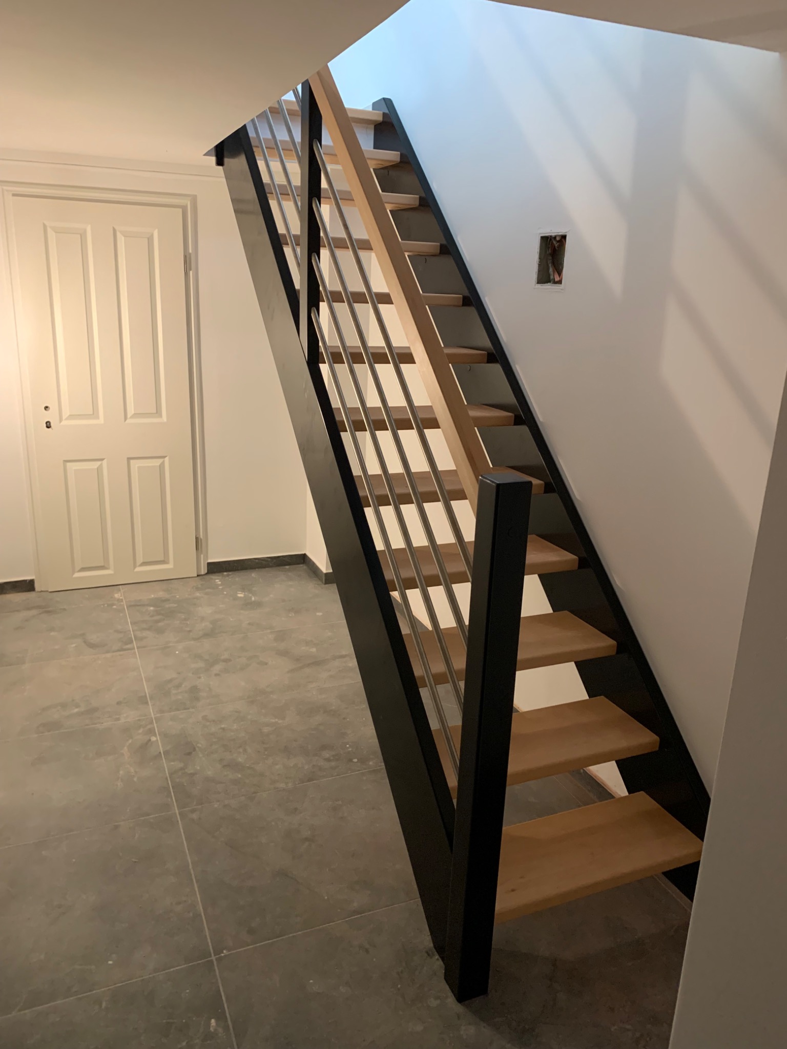 Nowoczesne schody z drewnianymi stopniami, czarną konstrukcją i metalowymi prętami balustrady, widoczne na tle białej ściany z gniazdkiem elektrycznym i białych drzwi.