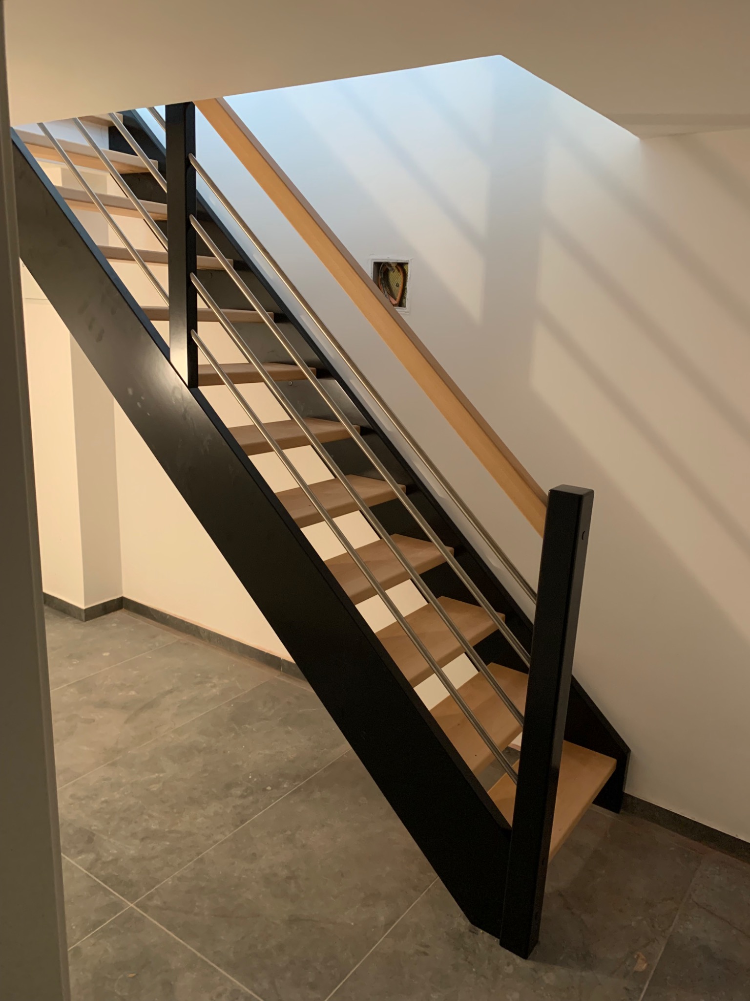 Nowoczesne schody z drewnianymi stopniami, czarną konstrukcją i metalowymi poręczami, widoczne z dołu w jasnym wnętrzu.