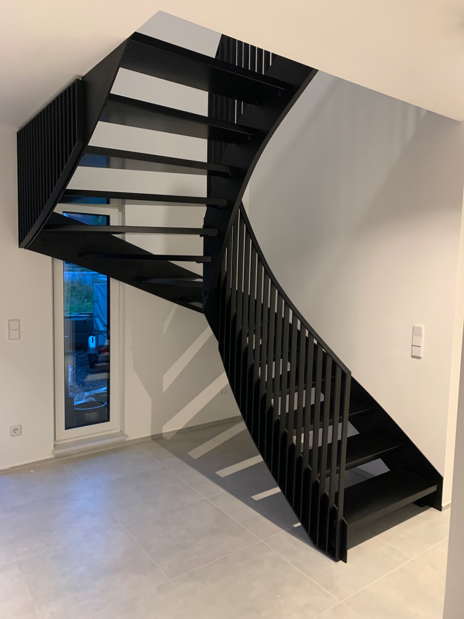 Nowoczesne, czarne, metalowe schody spiralne z minimalistyczną balustradą w jasnym wnętrzu z szarymi płytkami podłogowymi.