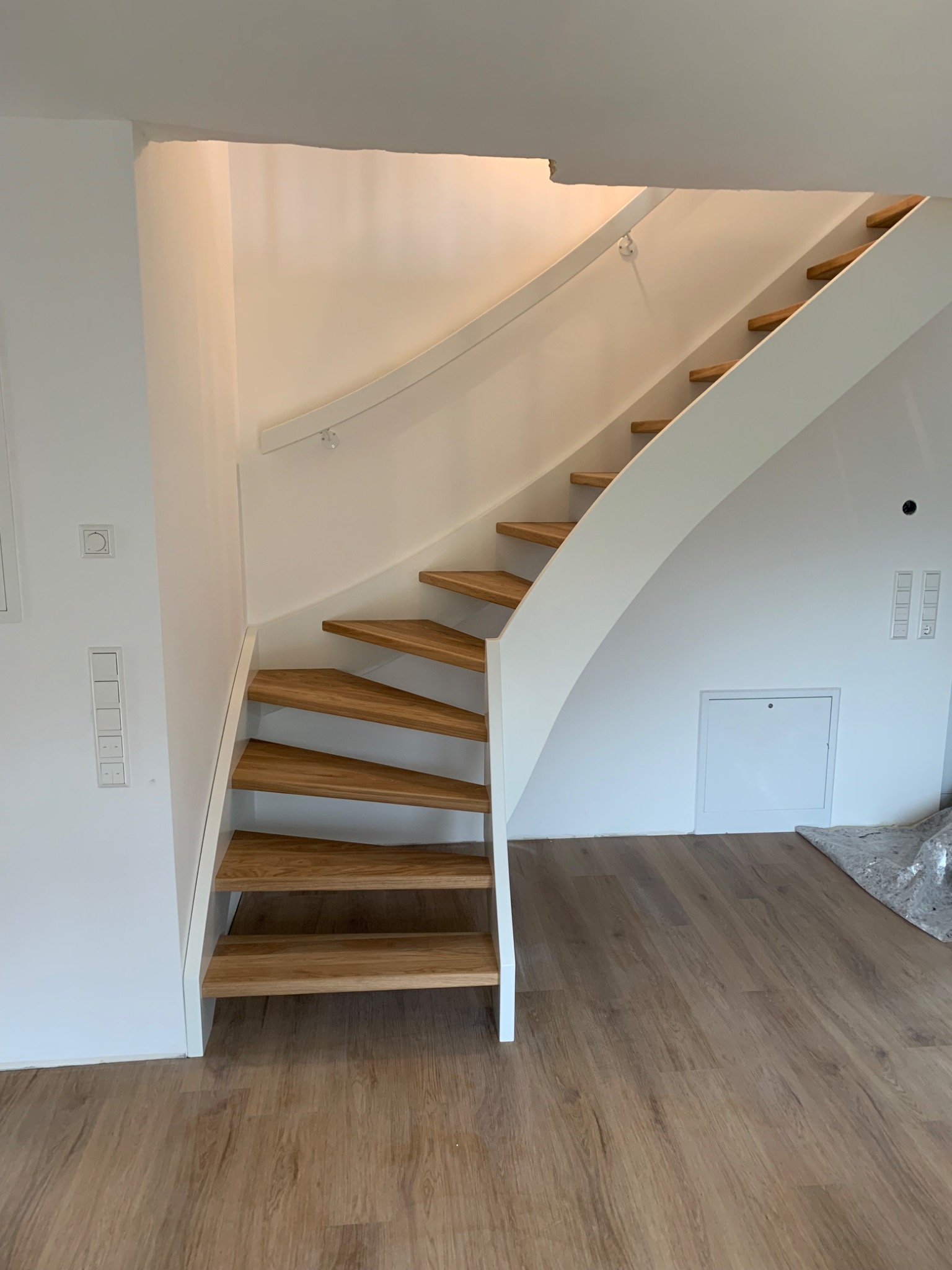 Nowoczesne schody z drewnianymi stopniami i białą, zakrzywioną konstrukcją, z delikatną poręczą, w jasnym wnętrzu z panelami podłogowymi.