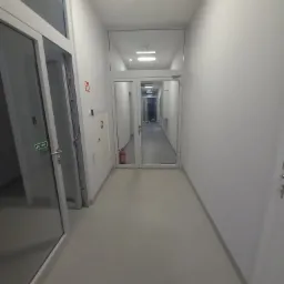 Korytarz biurowy w Ożarowie Mazowieckim z białymi ścianami, szarymi podłogami i przeszklonymi drzwiami na końcu, widoczny sprzęt przeciwpożarowy.