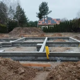 Wylane fundamenty domu z bloczków betonowych i wierzchnią warstwą betonu, widoczne żółte rury instalacyjne, w tle dom z panelami słonecznymi na dachu i sterta ziemi.