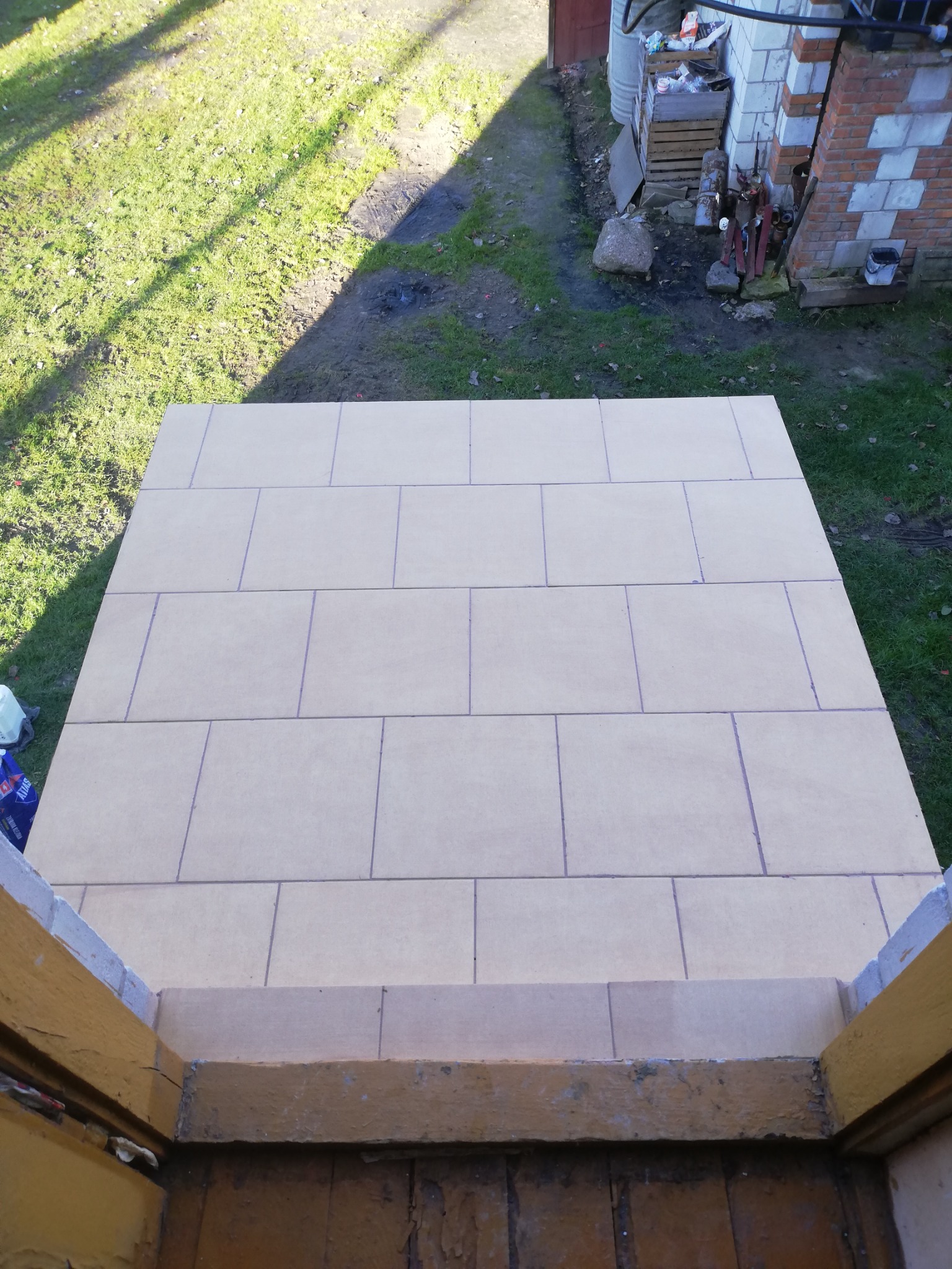 Widok z góry na płytki ułożone na zewnątrz, prawdopodobnie na tarasie lub schodach, z widocznymi fugami i elementami otoczenia, takimi jak trawa i budynek gospodarczy.