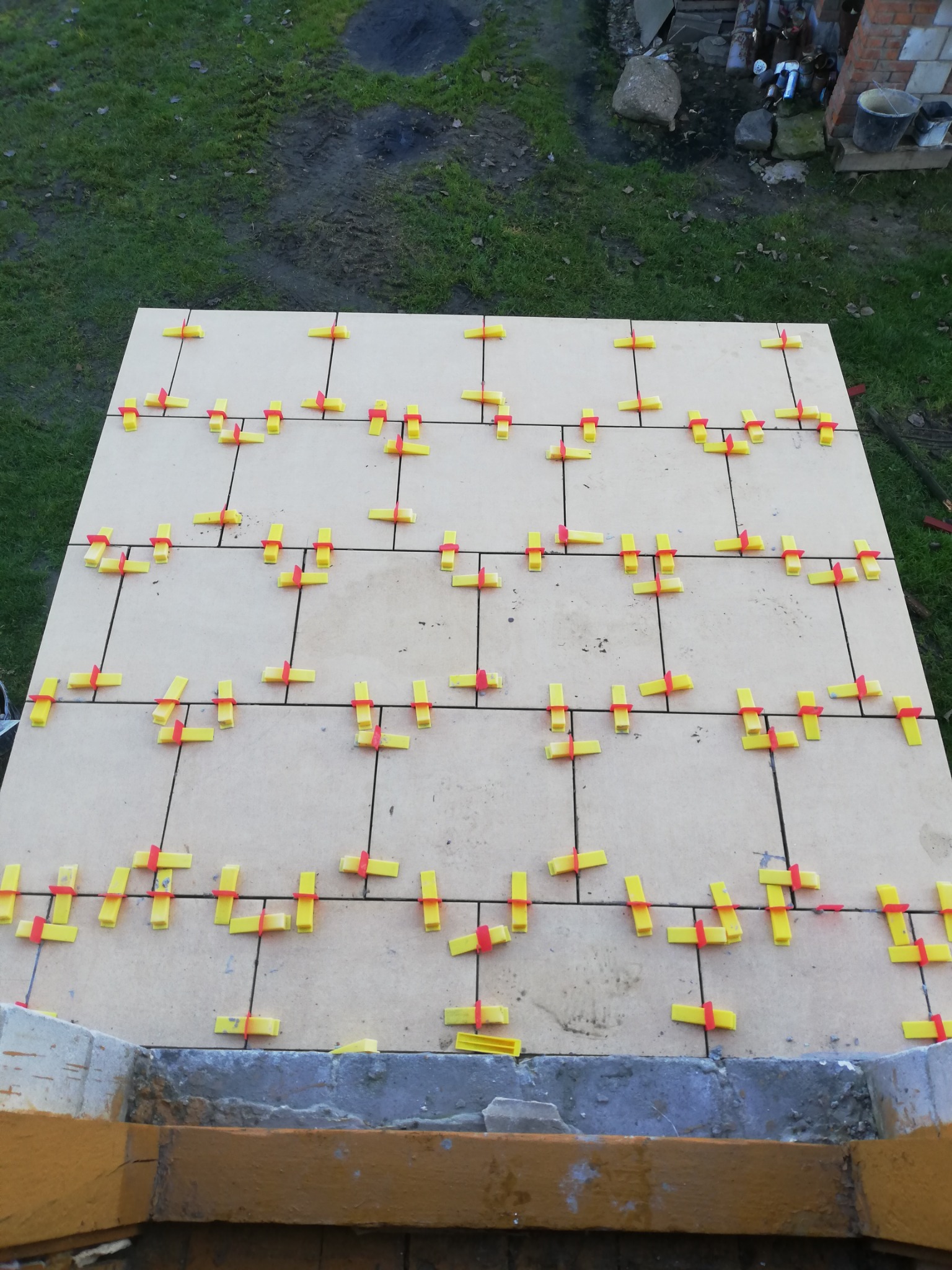 Układanie płytek na podłodze z użyciem żółto-czerwonych krzyżyków dystansowych, widok z góry na częściowo ukończoną powierzchnię.