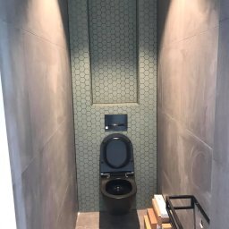 Buw-Koral Karol Brozdowski - Wąska toaleta z szarymi płytkami ściennymi i heksagonalnymi płytkami za wiszącą muszlą klozetową, podświetlona od góry, z kartonami i metalowym stelażem na podłodze.