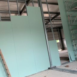 Buw-Koral Karol Brozdowski - Montaż ścian działowych z płyt gipsowo-kartonowych w hali przemysłowej, widoczne metalowe profile konstrukcyjne i drabina oparta o ścianę.