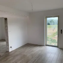 Wnętrze pokoju w trakcie remontu, widoczne ułożone panele podłogowe imitujące drewno, białe ściany, okno z widokiem na zewnątrz, fragment niedokończonego otworu drzwiowego z odsłoniętą cegłą...