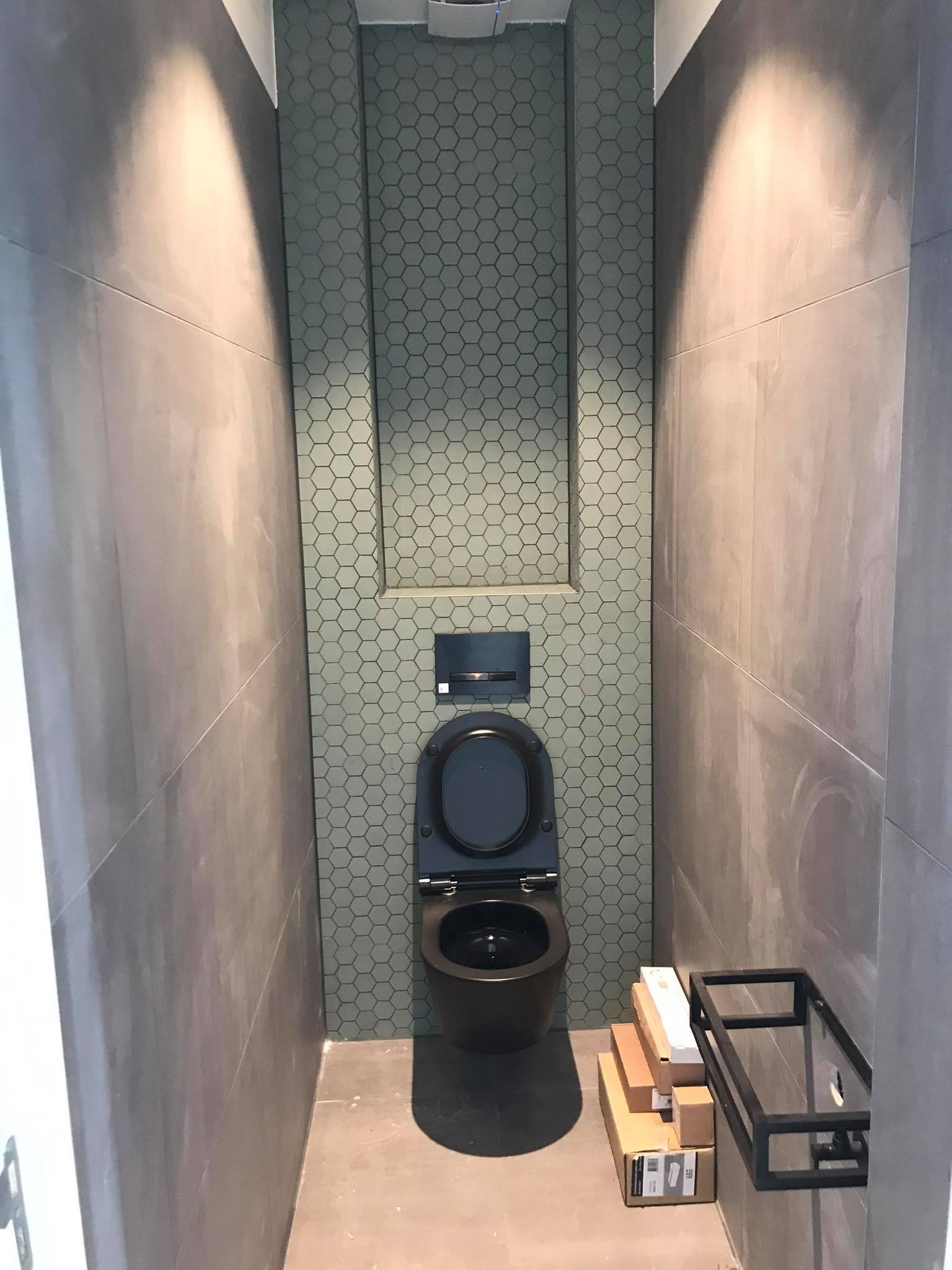 Wąska toaleta z szarymi płytkami ściennymi i heksagonalnymi płytkami za wiszącą muszlą klozetową, podświetlona od góry, z kartonami i metalowym stelażem na podłodze.