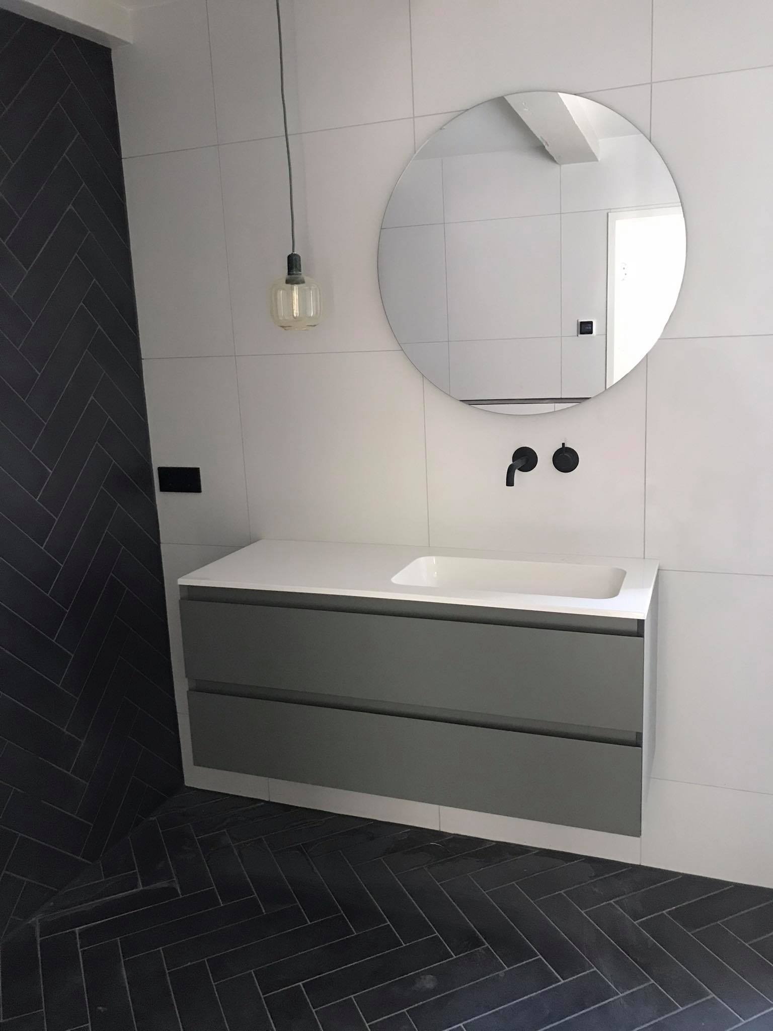 Nowoczesna łazienka z szarymi płytkami ułożonymi w jodełkę na podłodze i ścianie, białe płytki na pozostałych ścianach, okrągłe lustro nad szarą szafką z umywalką, czarna bateria i wisząca lampa...