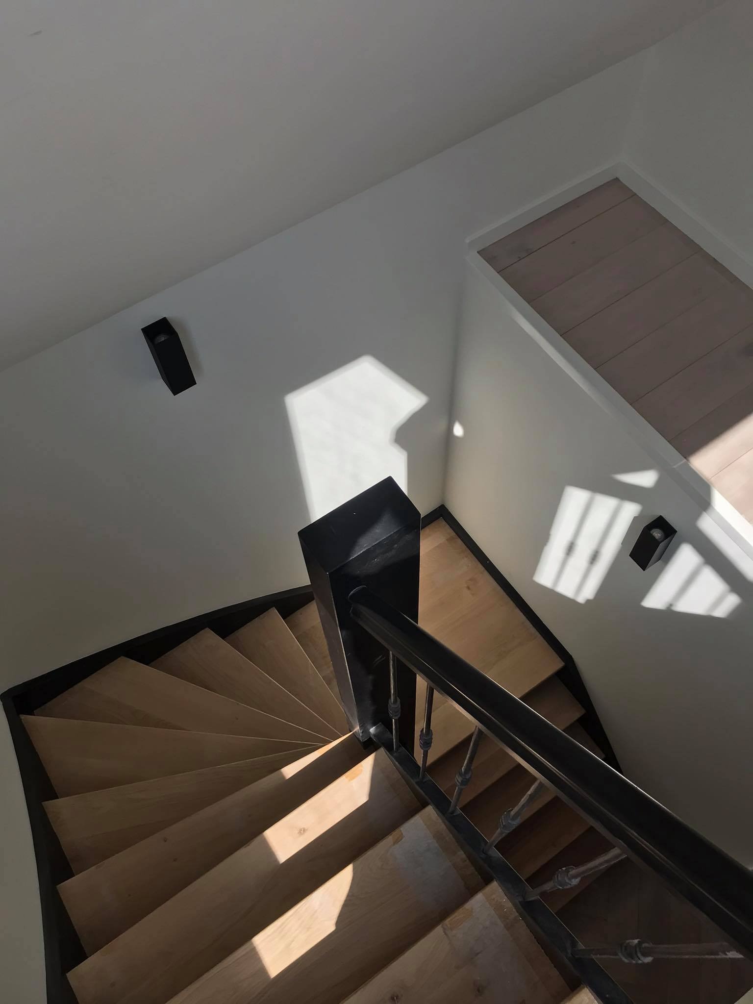Widok z góry na spiralne drewniane schody z czarną balustradą i oświetleniem ściennym, z promieniami słońca wpadającymi przez okno.