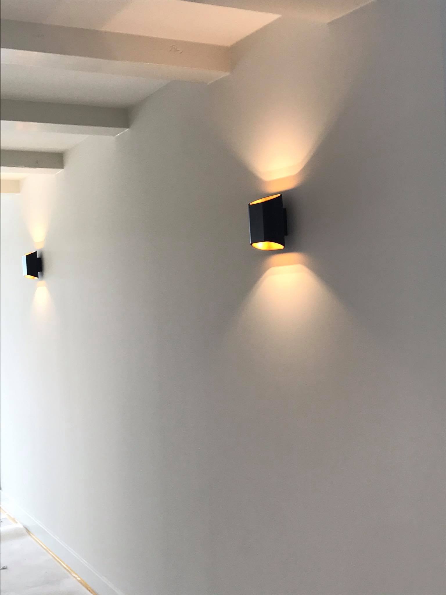 Dwa czarne, nowoczesne kinkiety ścienne emitujące ciepłe światło, zamontowane na gładkiej, białej ścianie w korytarzu z widocznymi elementami sufitu podwieszanego.