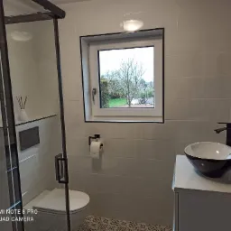 Nowoczesna łazienka z białymi płytkami, czarnymi akcentami, kabiną prysznicową typu walk-in, umywalką nablatową i widokiem na ogród przez okno.