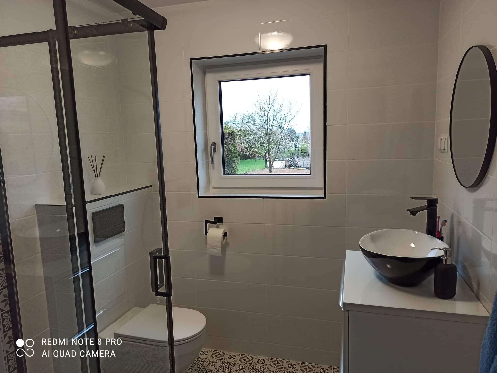 Nowoczesna łazienka z białymi płytkami, czarnymi akcentami, kabiną prysznicową typu walk-in, umywalką nablatową i widokiem na ogród przez okno.