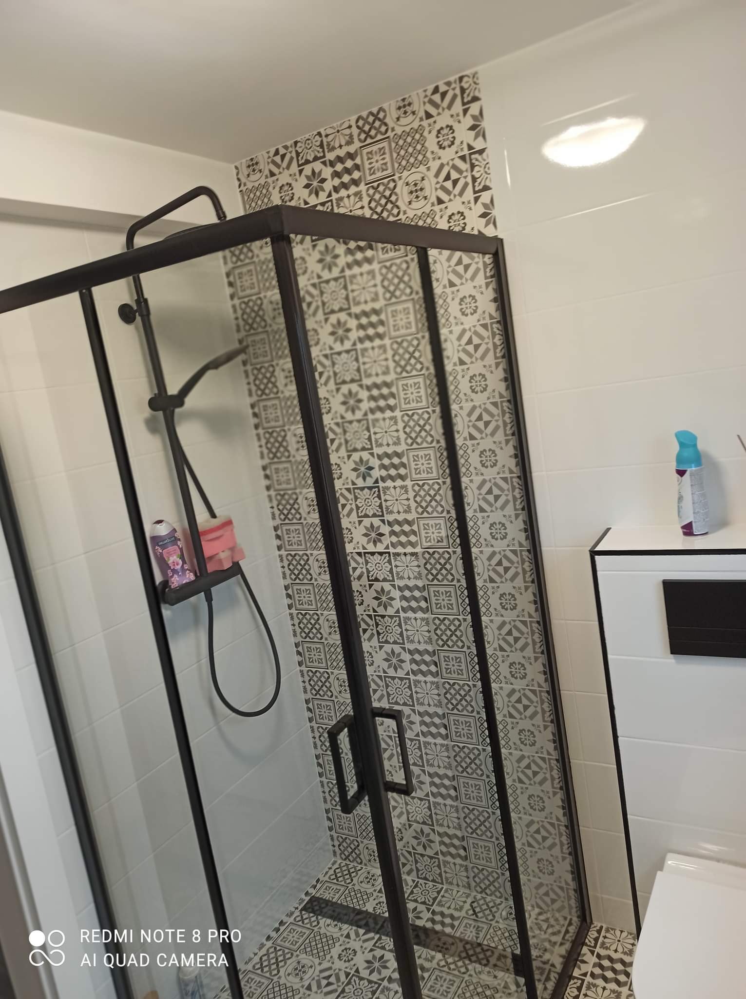 Kabina prysznicowa z czarnymi profilami i wzorzystymi płytkami na ścianach i podłodze, widoczny zestaw prysznicowy i kosmetyki na półce.