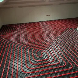 Czerwone rury ogrzewania podłogowego ułożone w spiralny wzór na czarnej macie izolacyjnej, widok z góry na poddaszu z oknem dachowym.