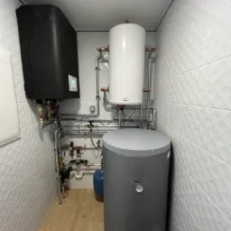 Kompaktowa kotłownia z szarym bojlerem, czarnym piecem dwufunkcyjnym, białym zbiornikiem wyrównawczym, instalacją rurową z zaworami i manometrami na tle białych płytek z delikatnym wzorem 3D...