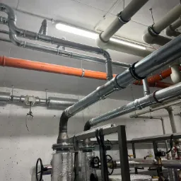Złożona instalacja rurowa z zaworami i izolacją, widoczne rury w różnych kolorach (szary, pomarańczowy) pod sufitem z oświetleniem jarzeniowym, elementy kotłowni w tle.