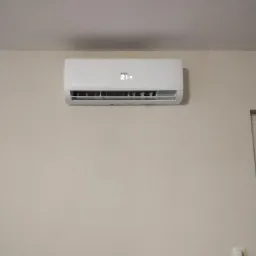 Biały klimatyzator marki Heiko zamontowany na beżowej ścianie, wyświetlający temperaturę 21 stopni Celsjusza, z widocznym włącznikiem światła w prawym dolnym rogu kadru.