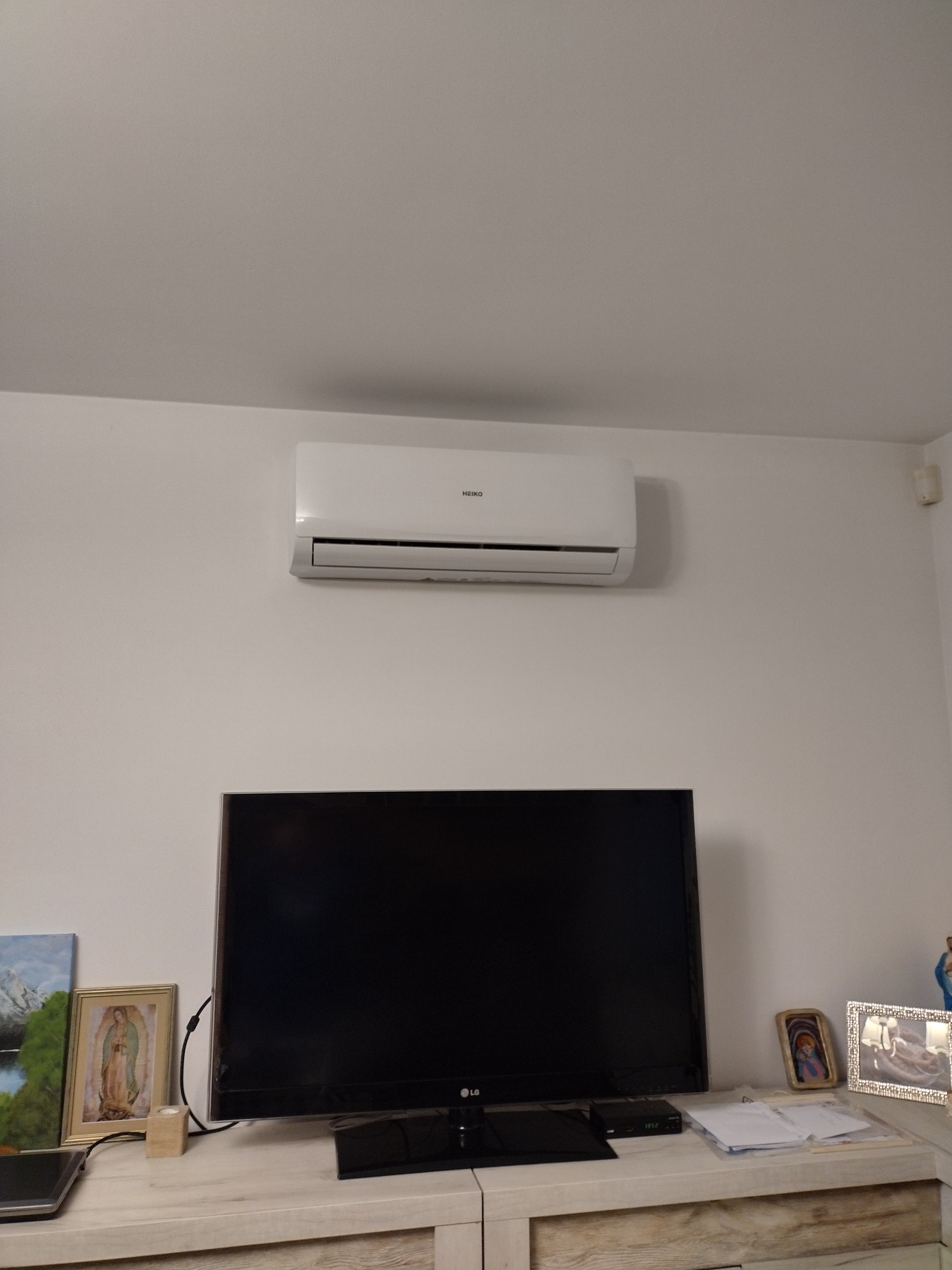 Biały klimatyzator marki HEIKO zamontowany na ścianie nad telewizorem w salonie. Widoczne dekoracje na szafce RTV.