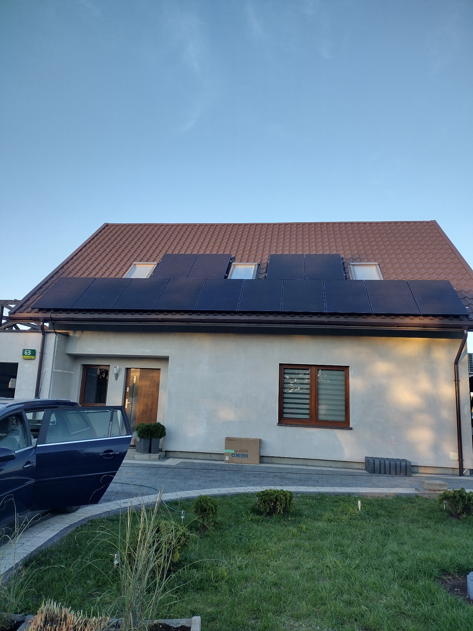 Dom jednorodzinny z brązowym dachem i panelami fotowoltaicznymi na połaci, widok z poziomu trawnika z zaparkowanym samochodem z otwartymi drzwiami i kartonem na ziemi.