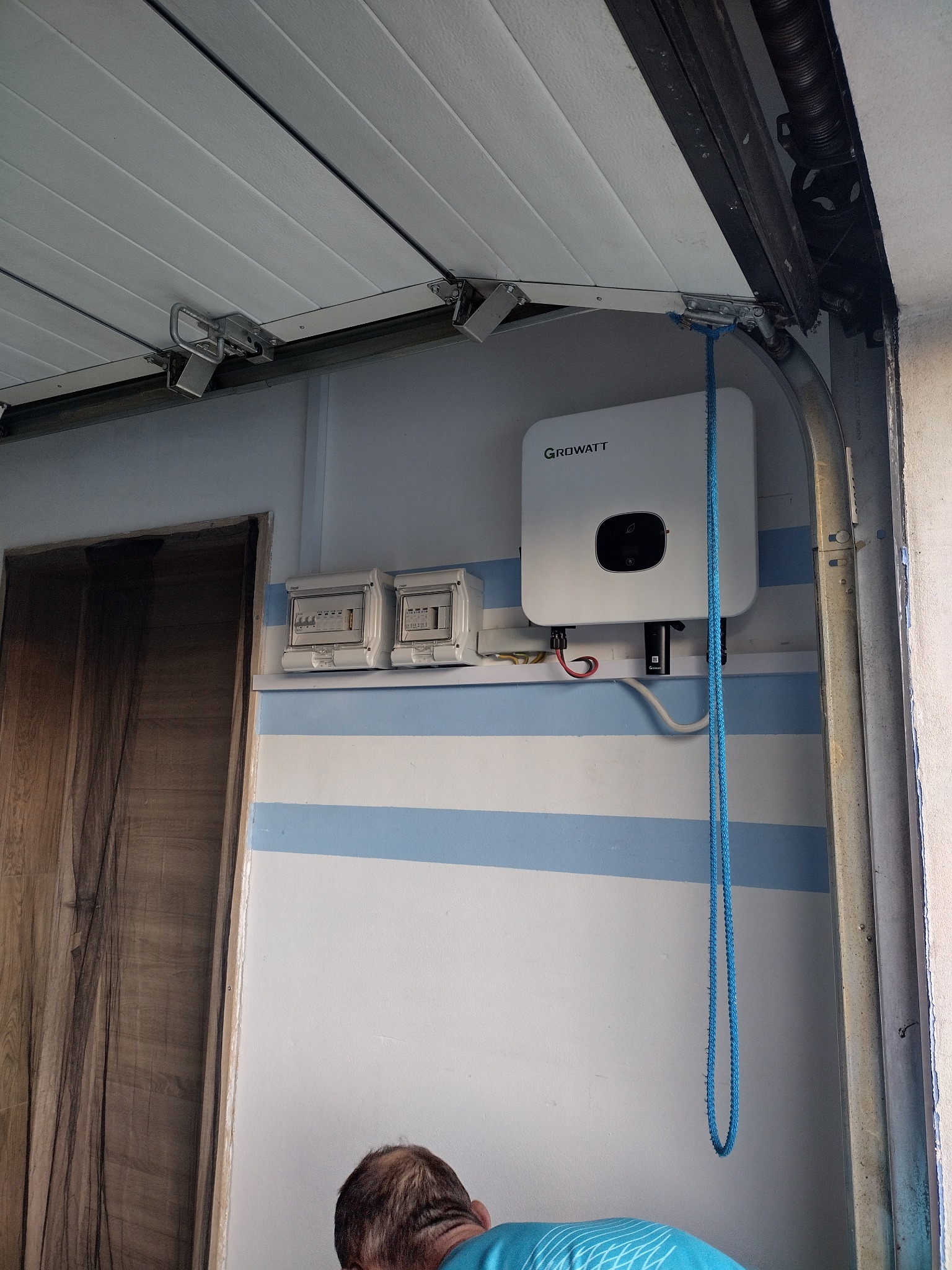 Zamontowany inwerter Growatt obok skrzynki bezpiecznikowej na ścianie garażu z niebieskimi pasami, widoczny fragment mechanizmu otwierania bramy garażowej i osoba w tle.