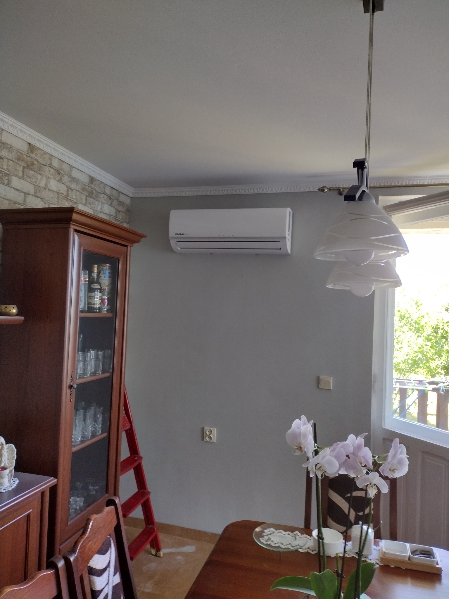 Wnętrze pokoju z zamontowanym klimatyzatorem Kaisai na szarej ścianie, widoczna również drewniana witryna, drabina, stół z orchideą i lampa sufitowa.