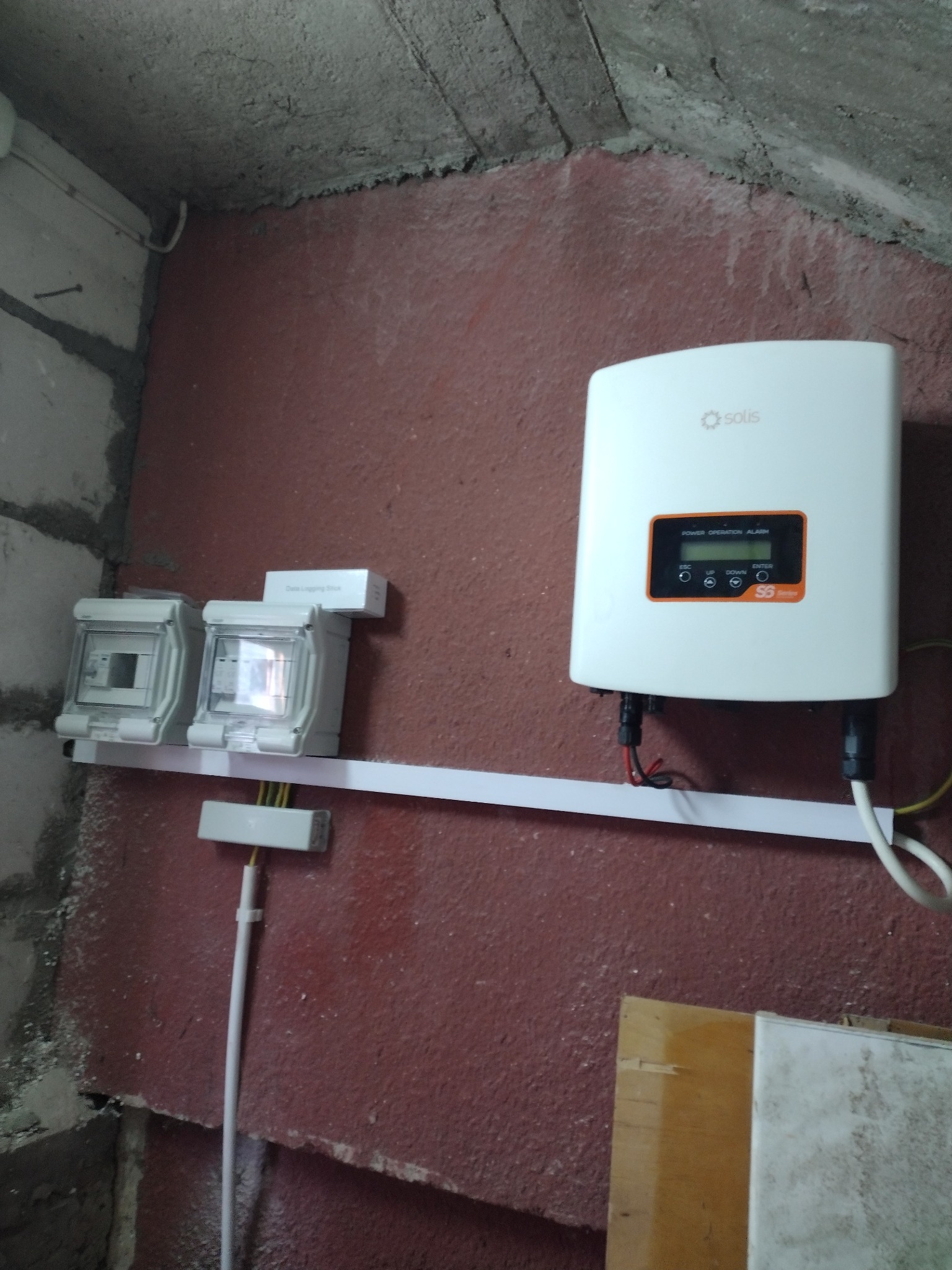 Biały falownik Solis zamontowany na ścianie z widocznymi zabezpieczeniami elektrycznymi i okablowaniem w surowym pomieszczeniu.