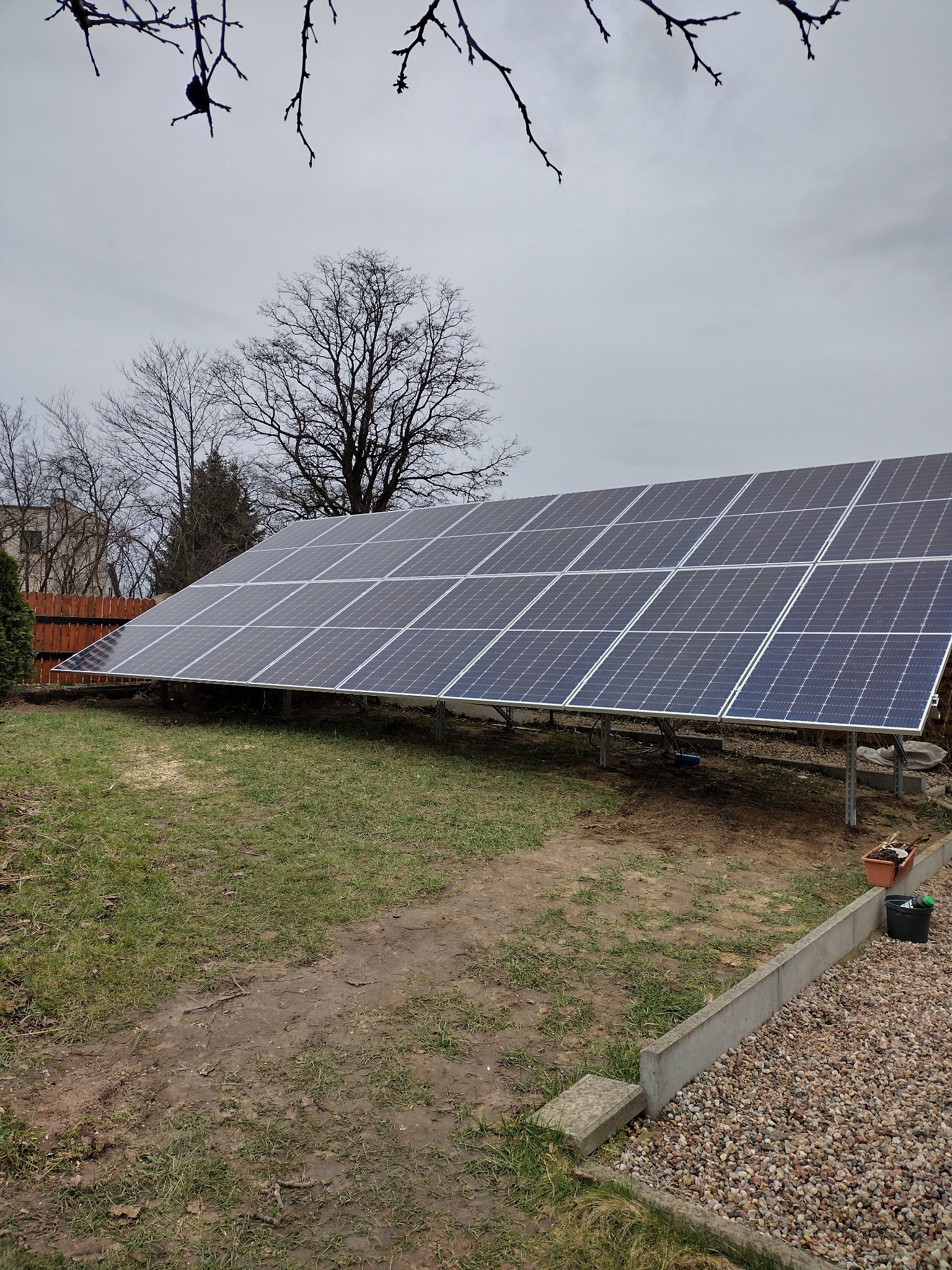 Panel fotowoltaiczny zamontowany w przydomowym ogródku na trawie, widoczne drzewa bez liści w tle i fragment drewnianego płotu.