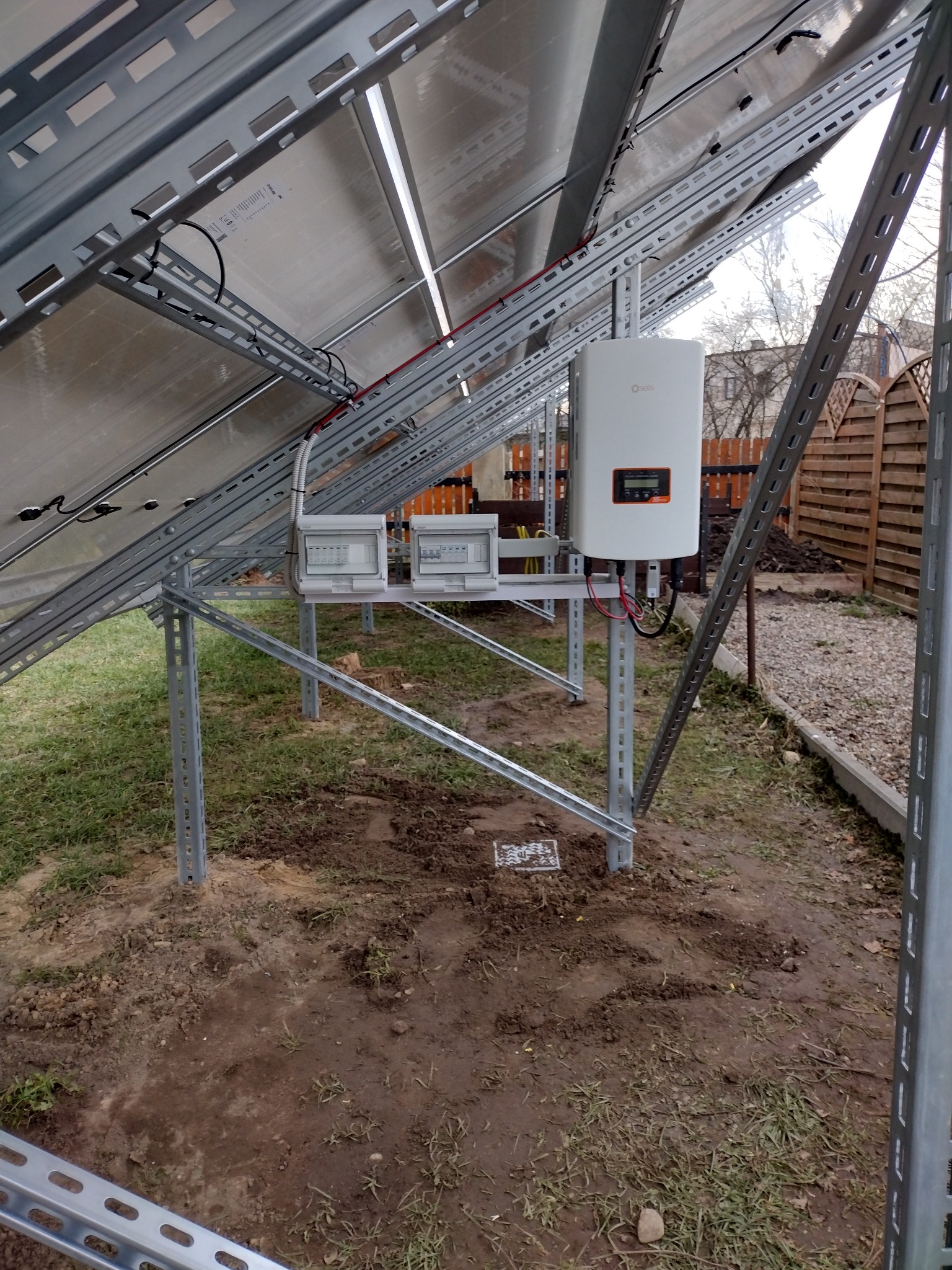 Widok od dołu na panele fotowoltaiczne zamontowane na metalowej konstrukcji, z widocznym inwerterem Solis i skrzynkami przyłączeniowymi, na tle ogrodu z drewnianym płotem.