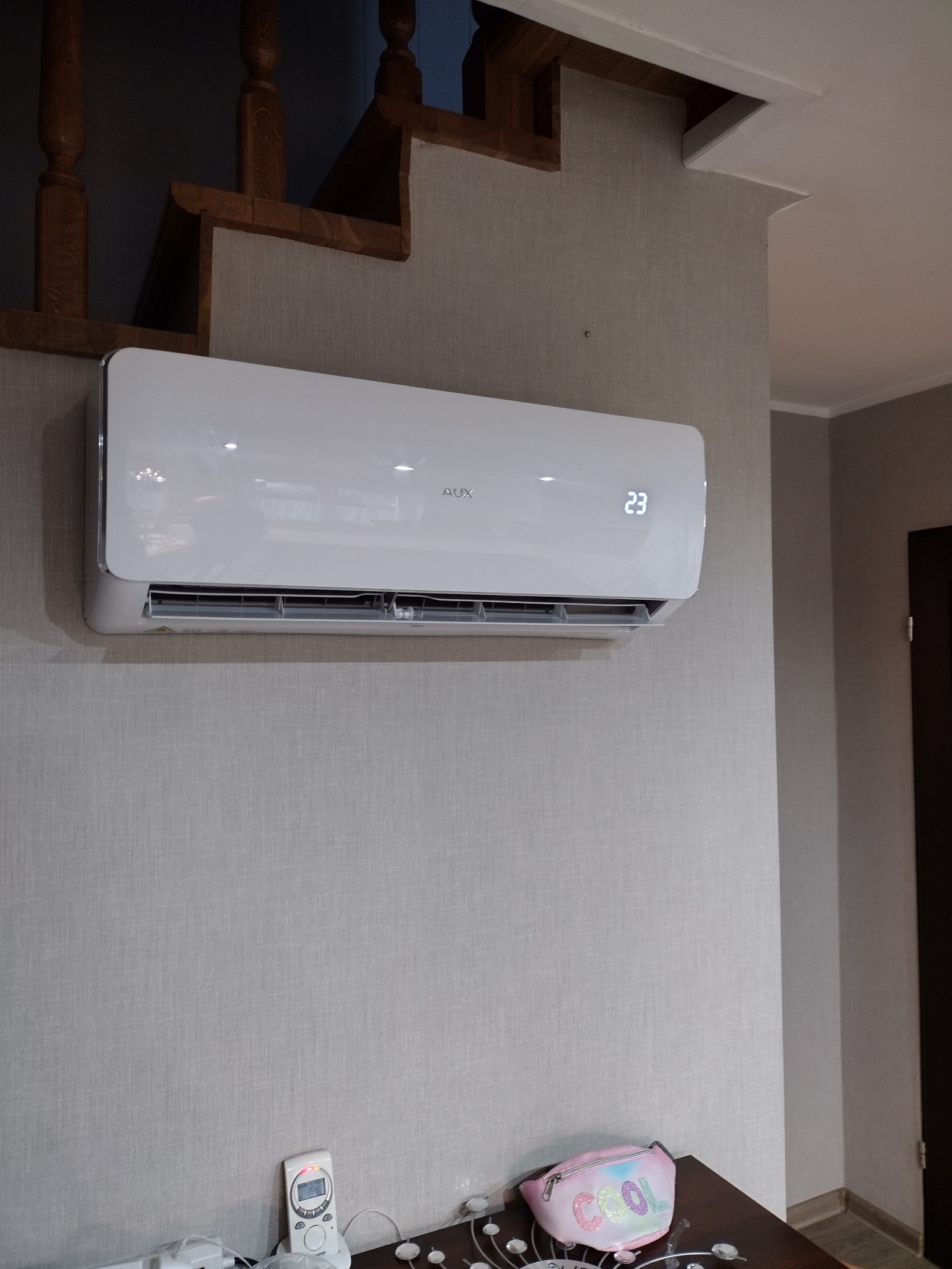 Biały klimatyzator AUX zamontowany na szarej ścianie z tapetą, widoczny fragment drewnianych schodów i wyświetlacz wskazujący 23 stopnie Celsjusza.
