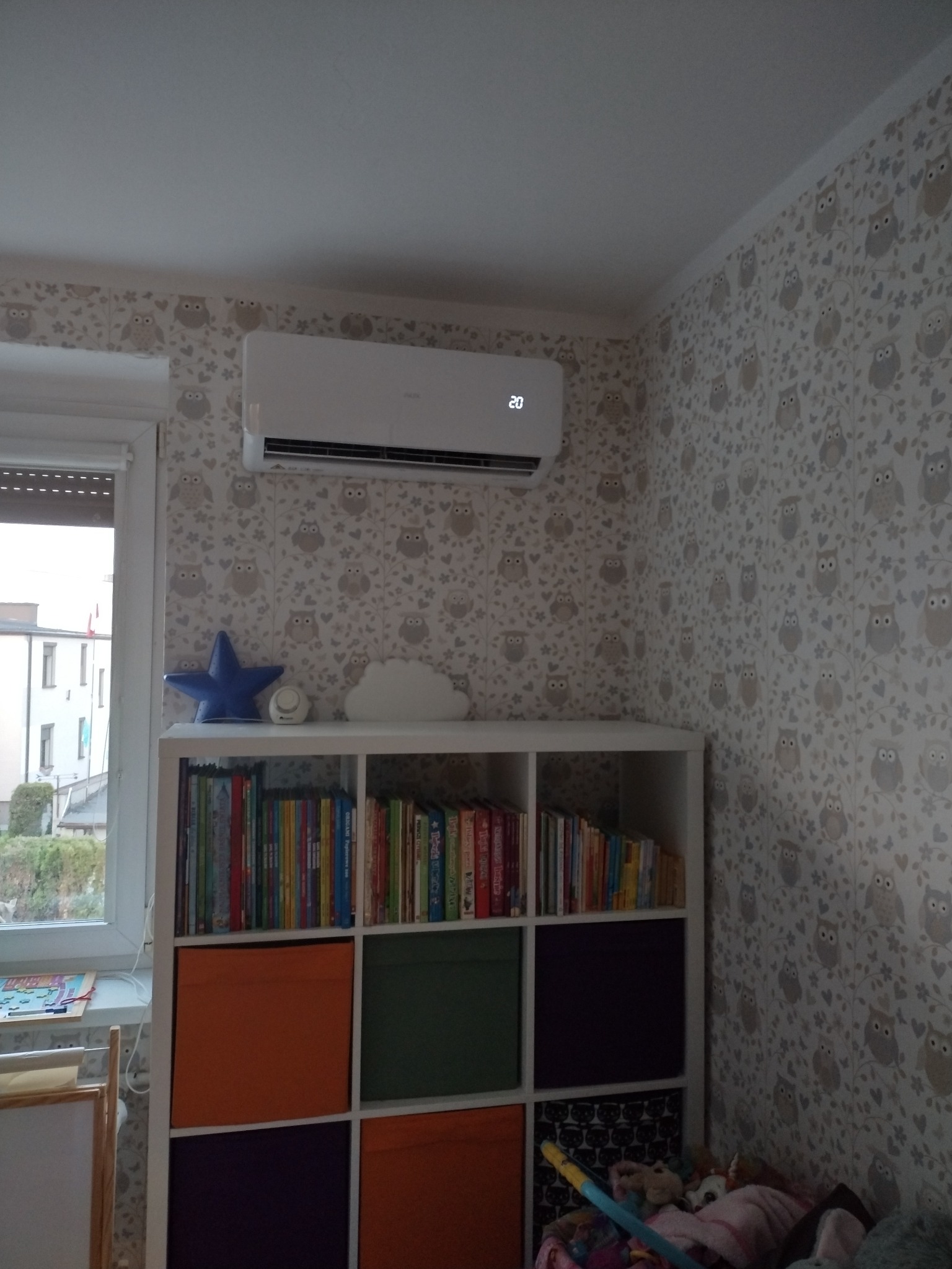 Biała jednostka wewnętrzna klimatyzatora na ścianie w pokoju dziecięcym z tapetą w sowy, wyświetlająca temperaturę 20 stopni, nad regałem z książkami i kolorowymi pojemnikami.