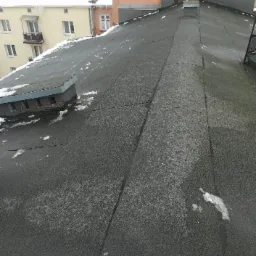 Czarny, płaski dach pokryty papą z niewielkimi pozostałościami śniegu, widoczny komin i fragmenty budynków w tle.