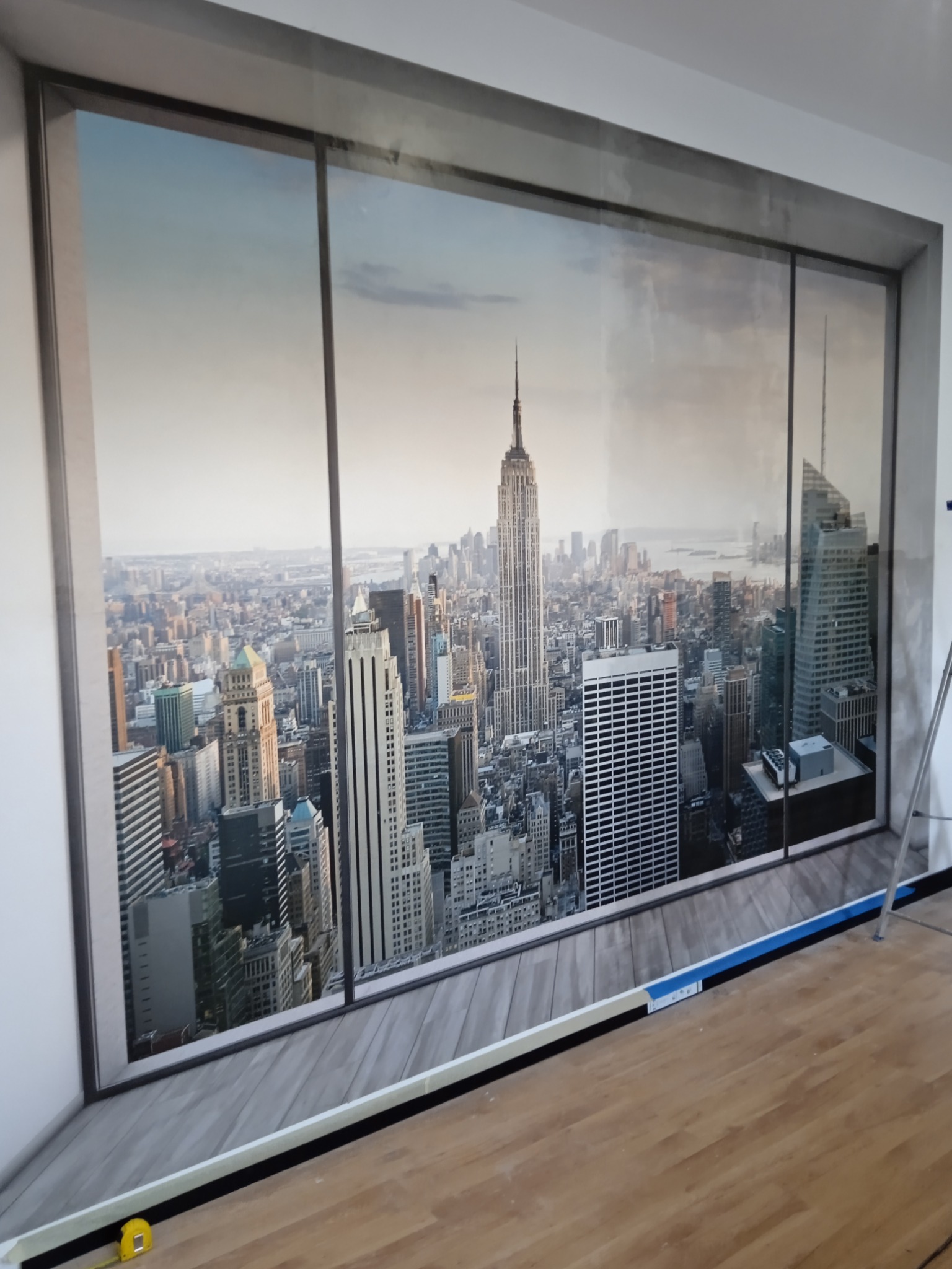 Fototapeta z panoramą Nowego Jorku, w tym Empire State Building, naklejona na ścianę w pomieszczeniu z drewnianą podłogą, widoczne narzędzia pomiarowe i statyw.