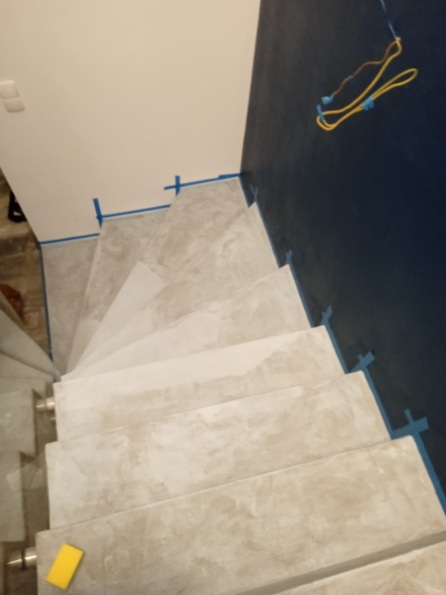 Schody wykończone w szarym mikrocemencie, zabezpieczone niebieską taśmą malarską, widok z góry, żółta gąbka leży na jednym ze stopni.