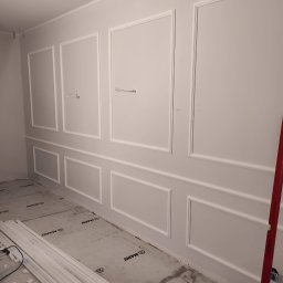 EMMAR MAREK WIŚNIEWSKI usługi remontowo budowlane - Ściana w trakcie dekorowania sztukaterią w postaci białych, prostokątnych paneli, widoczne przewody elektryczne, podłoga zabezpieczona folią, listwy przypodłogowe jeszcze nie zamontowane, poziomica...