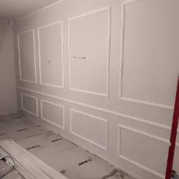 Ściana w trakcie dekorowania sztukaterią w postaci białych, prostokątnych paneli, widoczne przewody elektryczne, podłoga zabezpieczona folią, listwy przypodłogowe jeszcze nie zamontowane, poziomica...