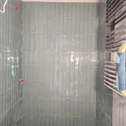 Pionowe, błyszczące płytki w odcieniach zieleni w trakcie układania w kabinie prysznicowej, widoczne krzyżyki dystansowe, drewnopodobna podłoga i grzejnik drabinkowy z boku.