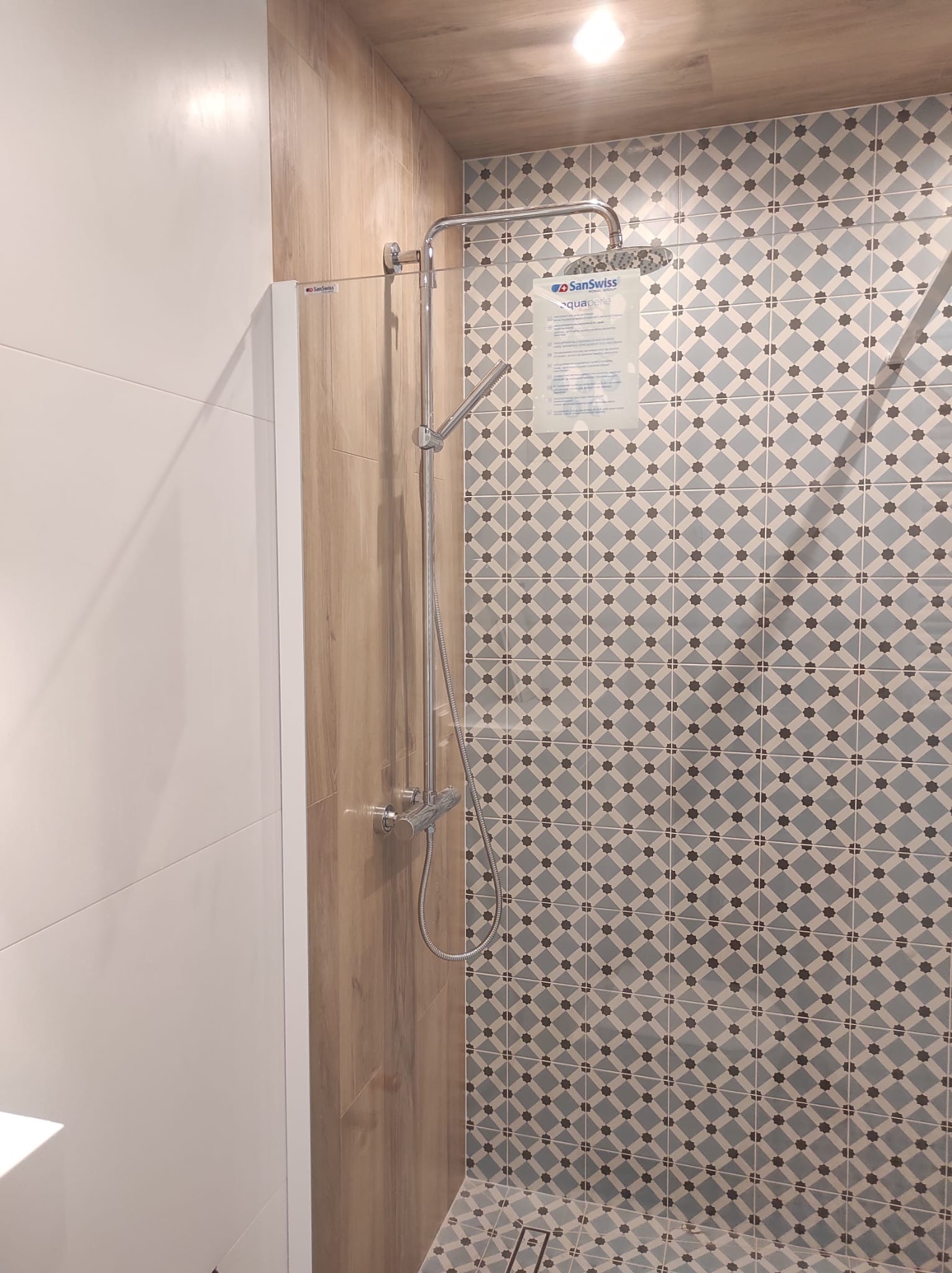 Kabina prysznicowa z deszczownicą, wykończona płytkami w geometryczny wzór, obok ściana z płytek imitujących drewno, na ścianie informacja o produkcie marki SanSwiss.