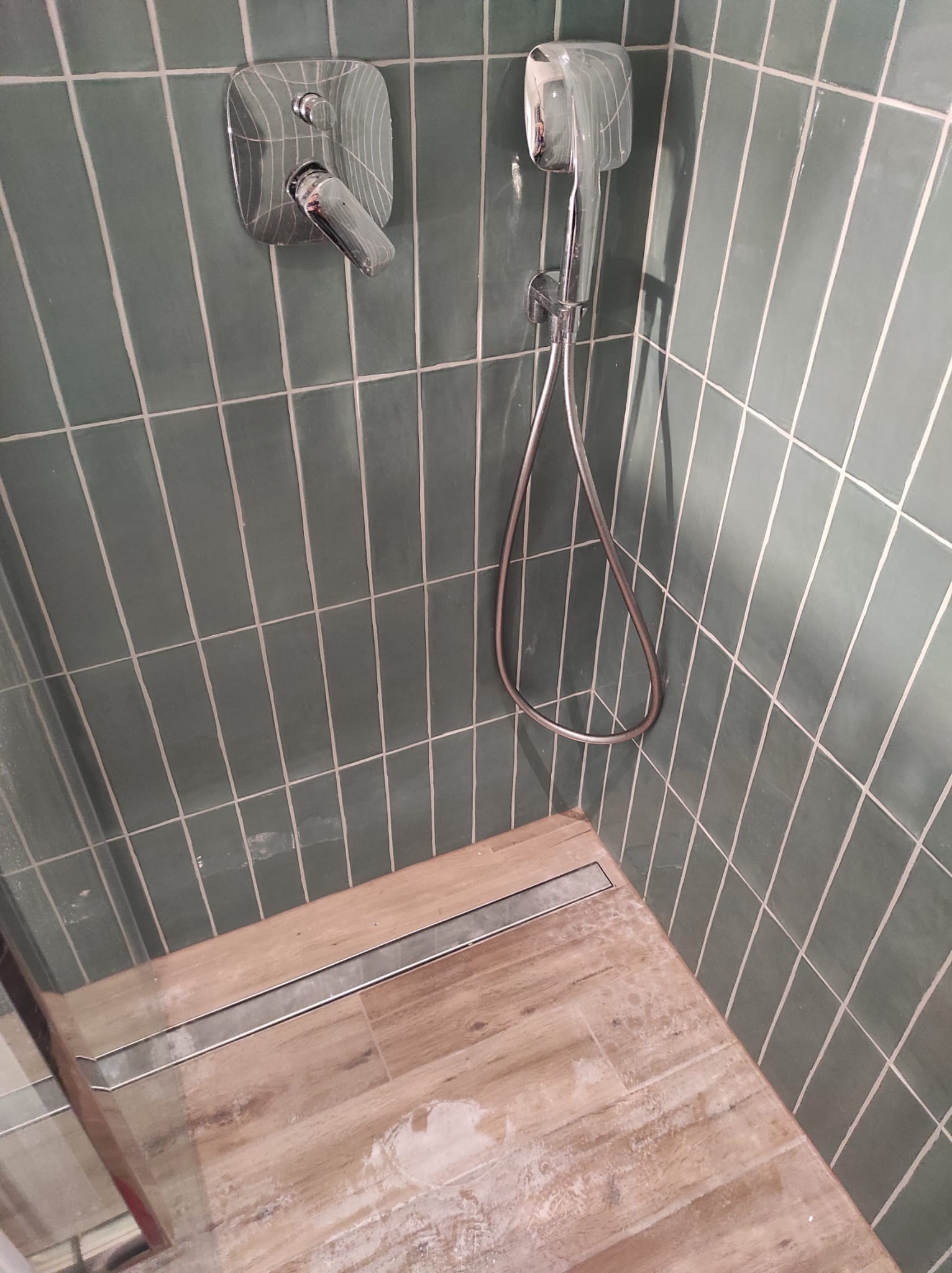 Kabina prysznicowa wyłożona pionowymi, oliwkowozielonymi płytkami z fugą, z zamontowaną armaturą, w tym słuchawką prysznicową, oraz podłogą imitującą drewno z odpływem liniowym.