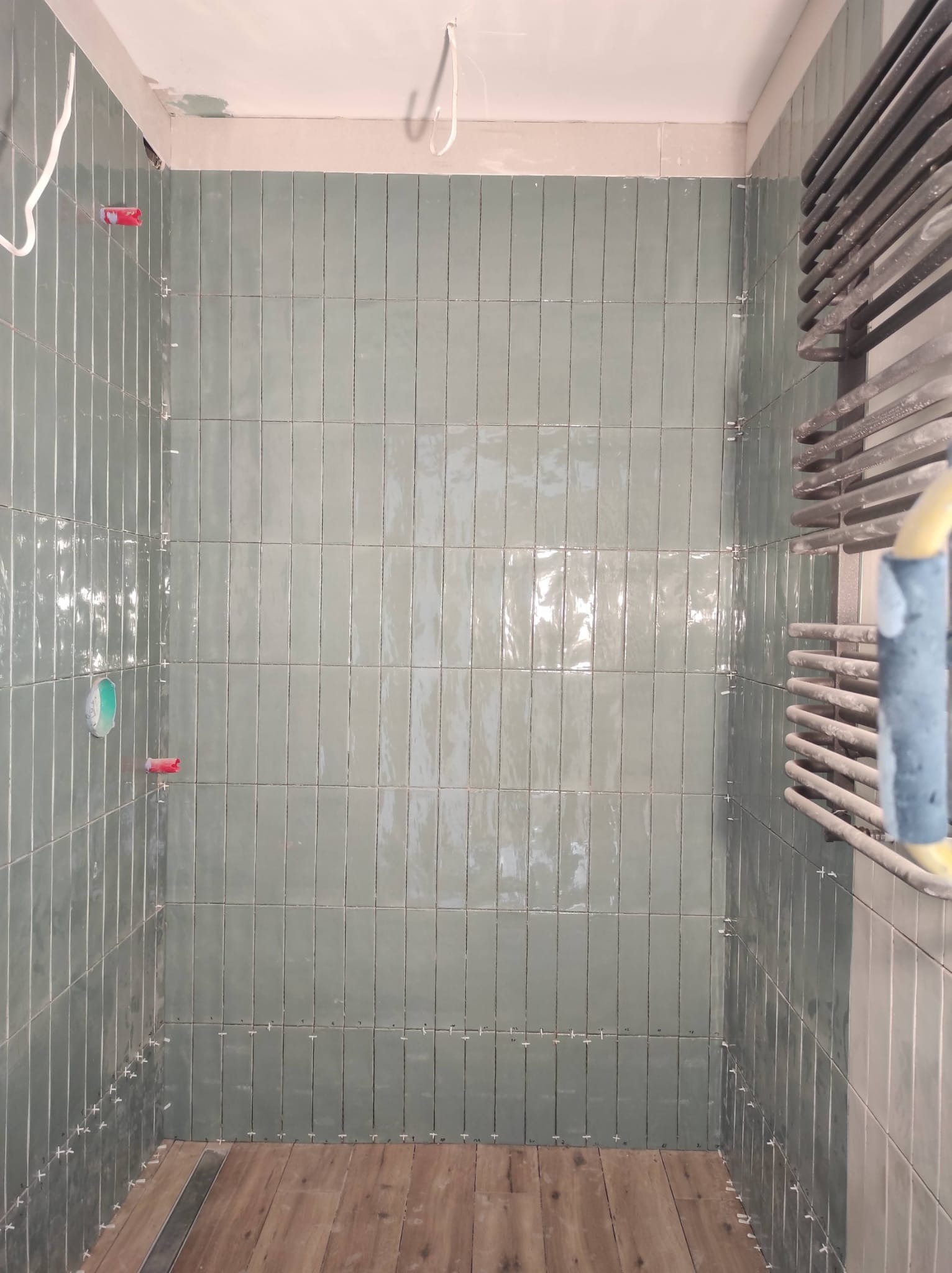 Pionowe, błyszczące płytki w odcieniach zieleni w trakcie układania w kabinie prysznicowej, widoczne krzyżyki dystansowe, drewnopodobna podłoga i grzejnik drabinkowy z boku.