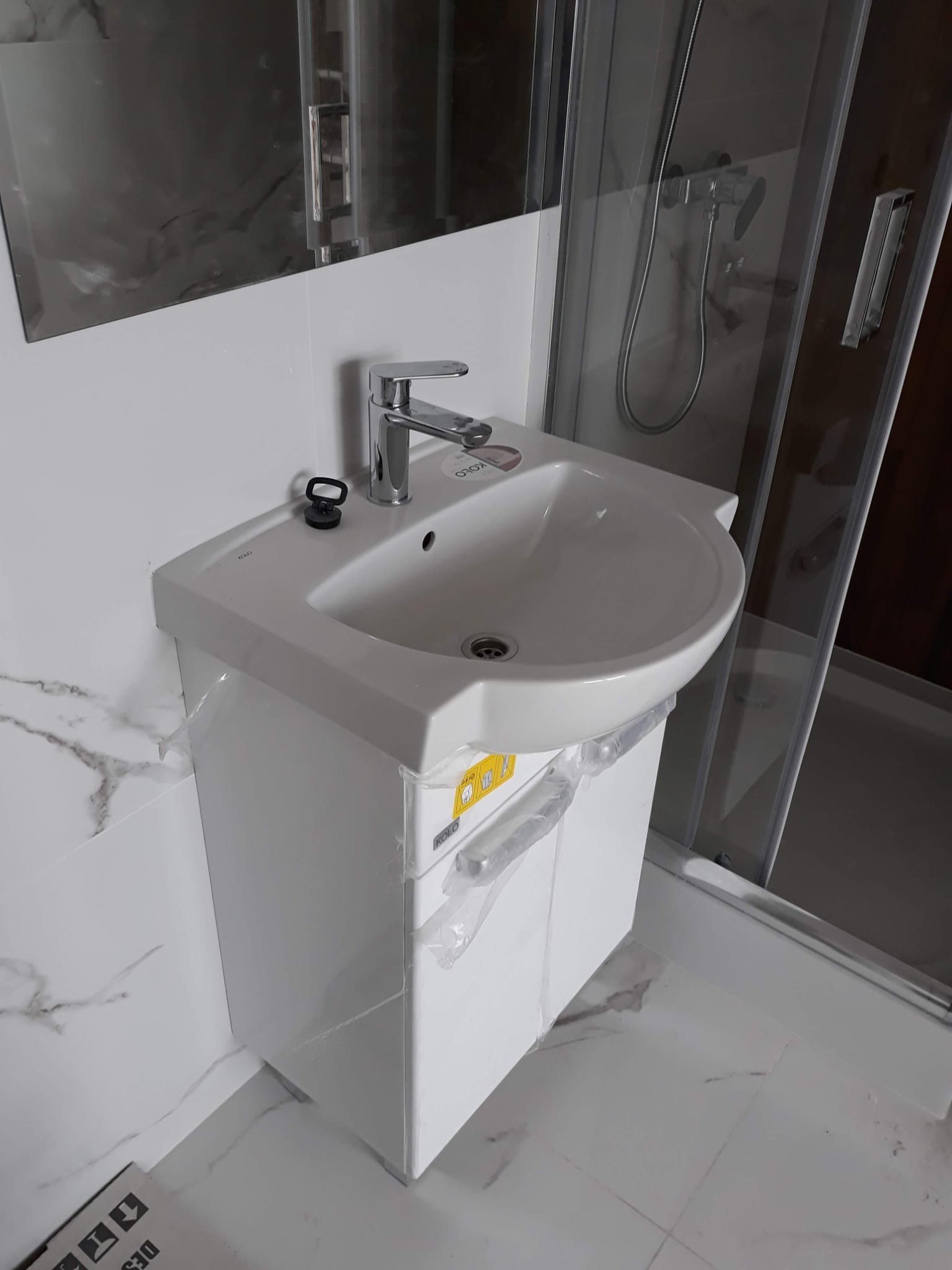Biała umywalka z szafką, model Koło, z chromowaną baterią, zamontowana w łazience z białymi płytkami imitującymi marmur, obok kabina prysznicowa ze srebrnymi profilami, widoczne folie ochronne...