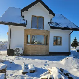 Nowoczesny dom jednorodzinny w zimowej scenerii, z białymi ścianami, drewnianą stolarką okienną i fragmentem elewacji wykończonym płytkami imitującymi drewno, klimatyzator na zewnątrz, stosy...